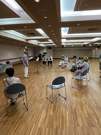 円形に配置した椅子に座る参加者、その中心で歌詞カードを持つ五城たかしさん