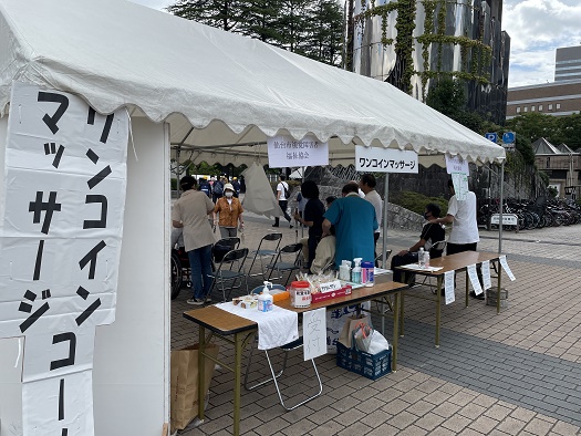 テントに仙台市視覚障害者福祉協会・ワンコインマッサージコーナーの看板、手前に受付のテーブル、奥に椅子に座って施術を受ける来場者と施術するマッサージ師