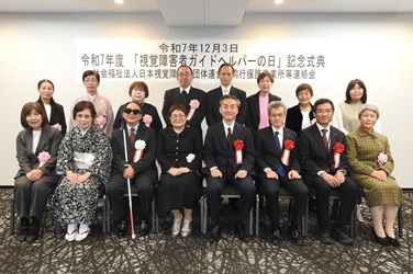 令和7年12月3日、令和7年度「視覚障害者ガイドヘルパーの日」記念式典の横断幕の前で前後16人の皆さんの記念写真