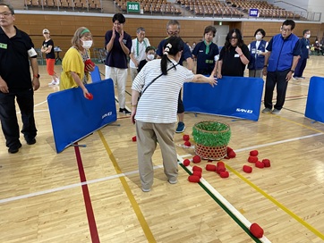 床に置かれた籠に玉を投げる女性参加者、隣に応援するチームメイト