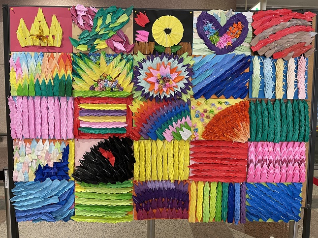 白いアートボードに貼り付けられた5×5で計25枚の色とりどりの折り鶴の作品